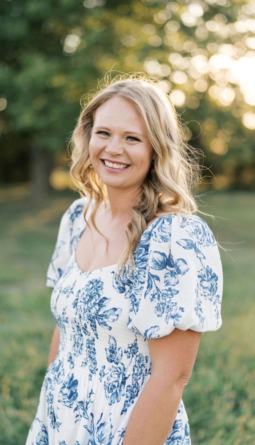 Becca Monroe outdoors in a white floral dress, smiling, with green trees and sunlight behind her